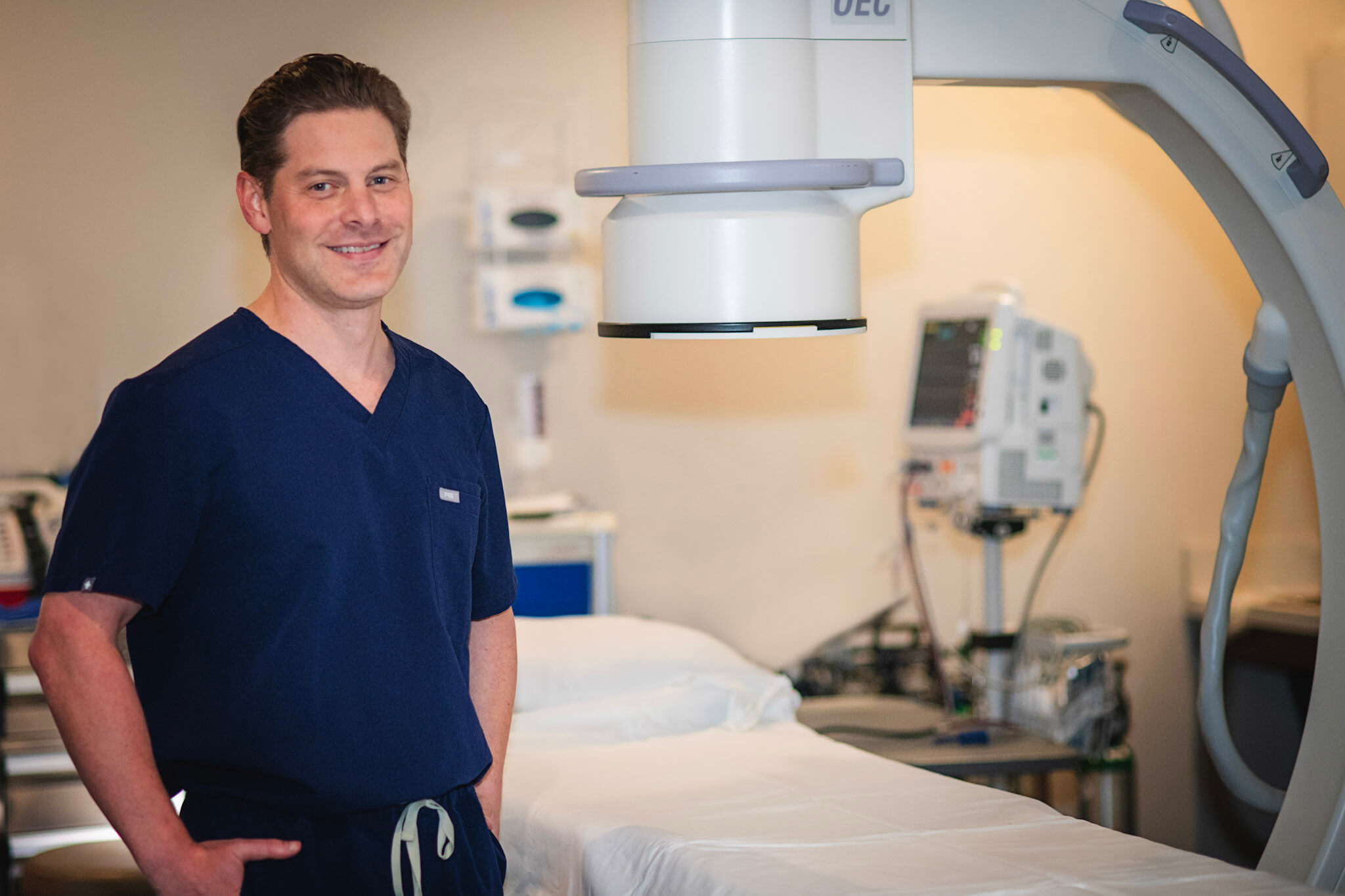 Dr. Johnson wearing dark blue scrubs standing beside a C-arm medical imaging machine